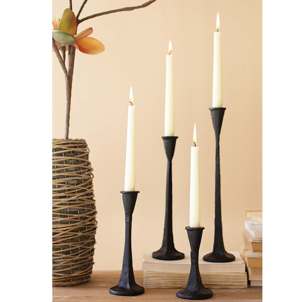 A set of black cast iron taper candle holders of varying heights, with white candles on top, placed on a wooden surface with a decorative item in the background.